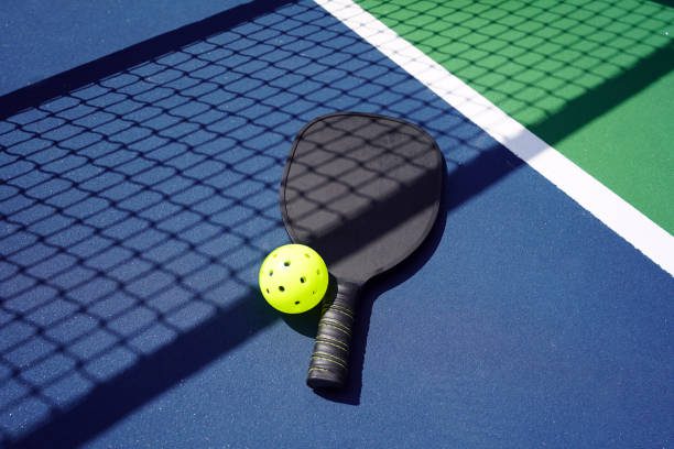 Pickle ball paddle with ball on court with net shadow.